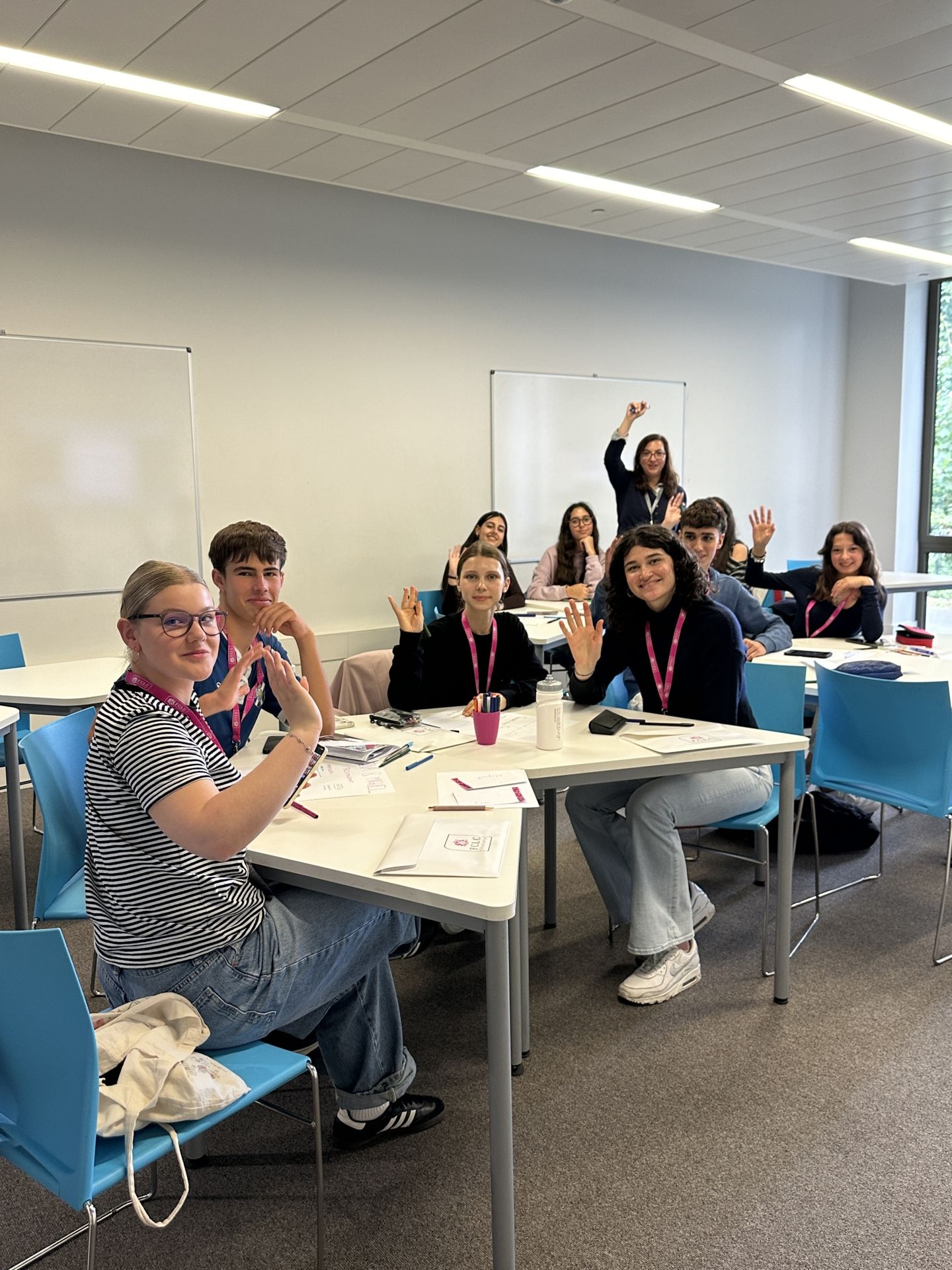 Image of 9 students and a teacher in a classroom waving at the camera